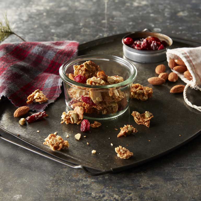 A glass jar of Morning Summit cereal with a small bag of almonds and a ramekin of cranberries